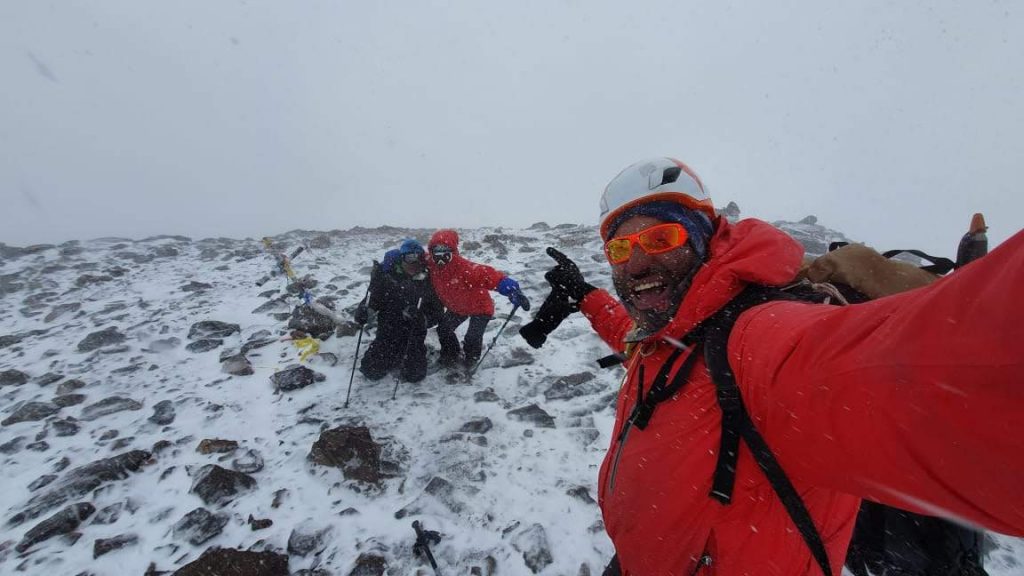 Para cégo ver: A foto é uma selfie e conta com três montanhistas em meio a uma nevasca fraca. O montanhista que está tirando a foto está dando risada e apontando para os dois colegas que estão ao fundo.