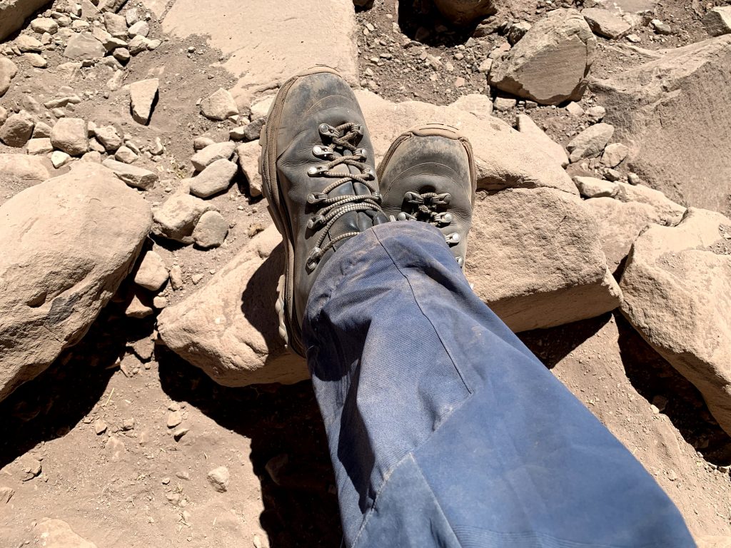 Para cégo ver: o montanhista tirou uma foto dos seus pés cruzados enquanto estava sentado. Ele está sobre rochas e pedregulhos. 