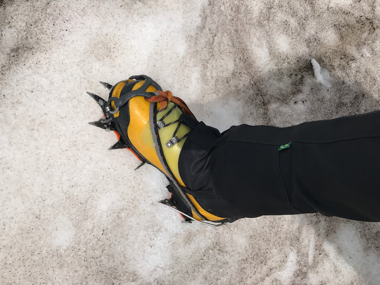Para cégo ver: o montanhista tirou uma foto apenas do seu pé direito calçado com uma bota e um crampon. Ele está sobre a neve.
